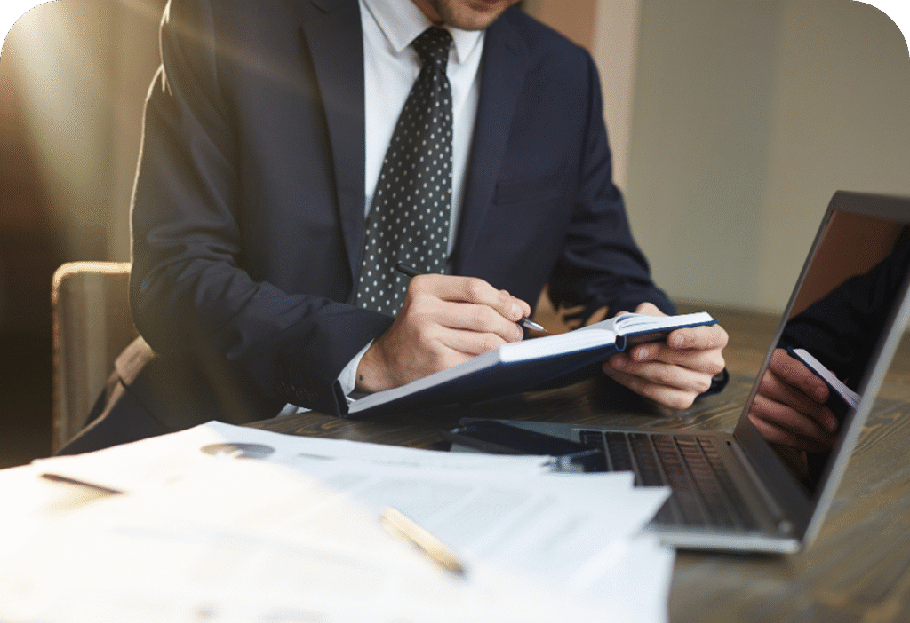 Man in suit writing in notebook at desk, with open laptop and papers scattered around.