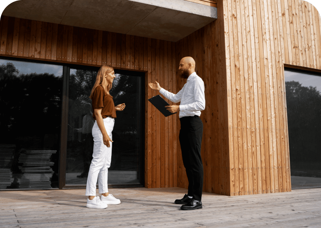 Two people talking outside a wooden building; one holds a folder.