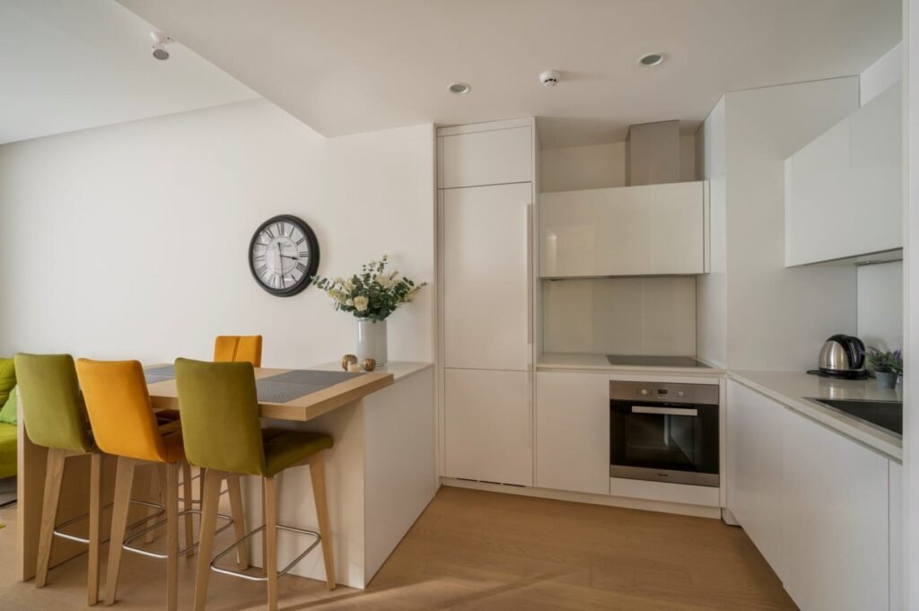 Modern kitchen with white cabinets, oven, kettle, and a dining area with colorful chairs. Wall clock and vase on counter.