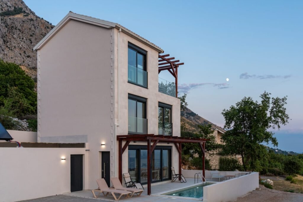 Three-storey villa under construction with glass balconies, pool, and sun loungers, set against a hillside with trees.